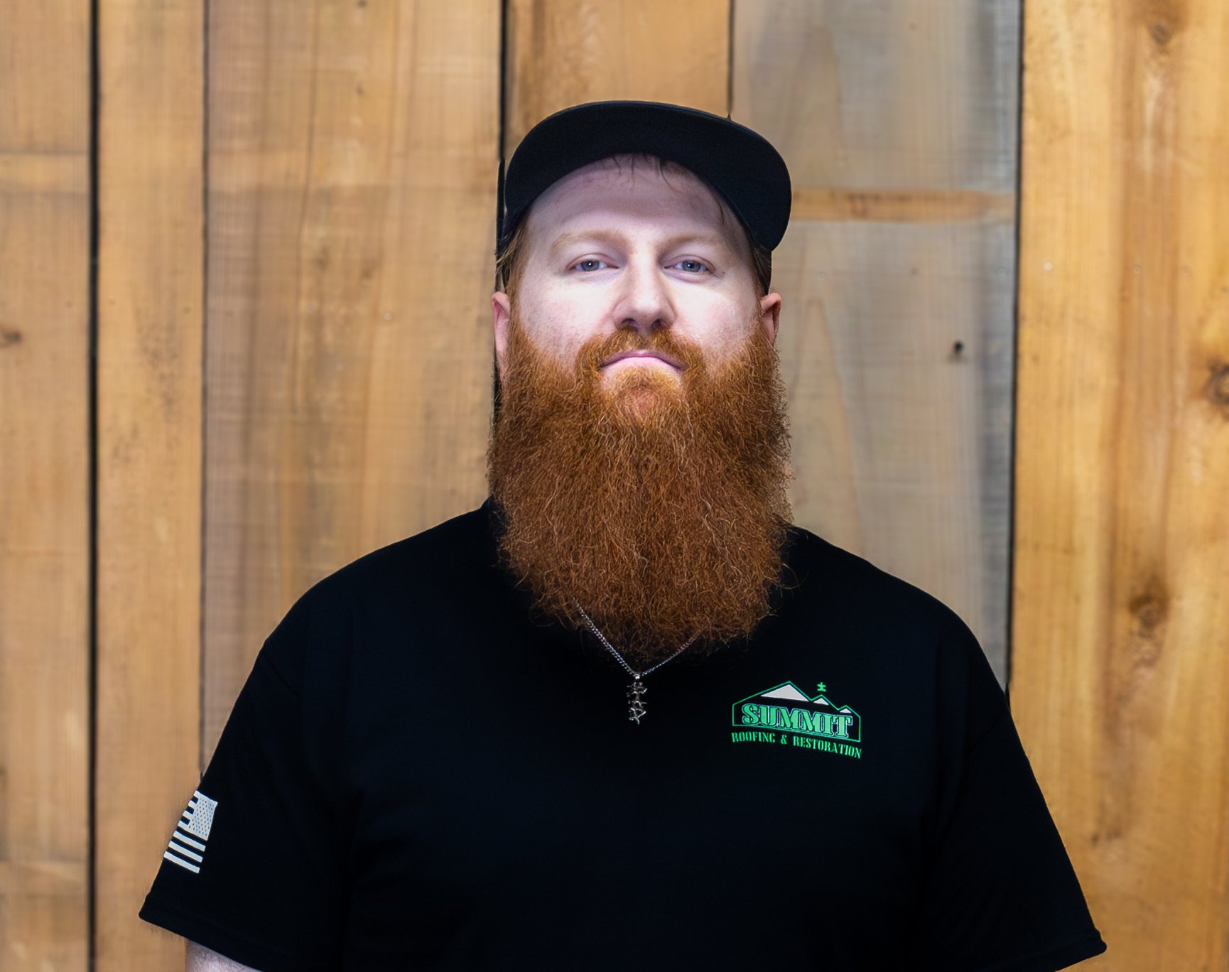 A bearded man wearing a black cap and a black Summit T-shirt stands in front of a wooden wall, looking directly at the camera with a neutral expression.