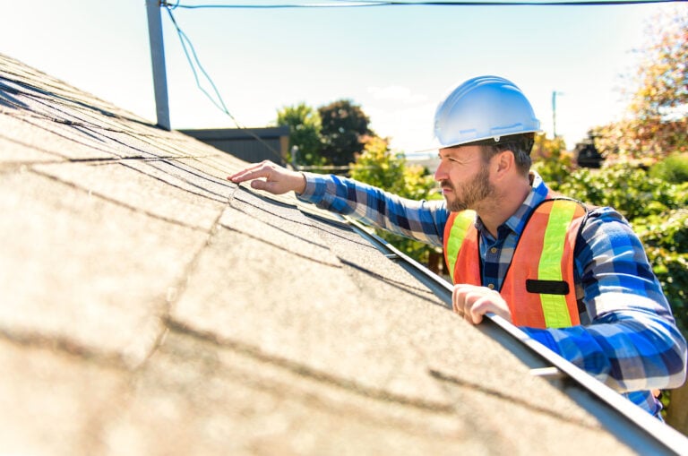 A construction worker in a blue hard hat and orange safety vest inspects a shingled roof, mindful of the roof inspection cost. He steadies himself on a ladder with one hand, closely examining the shingles with the other. It's sunny, with trees and power lines framing the scene.
