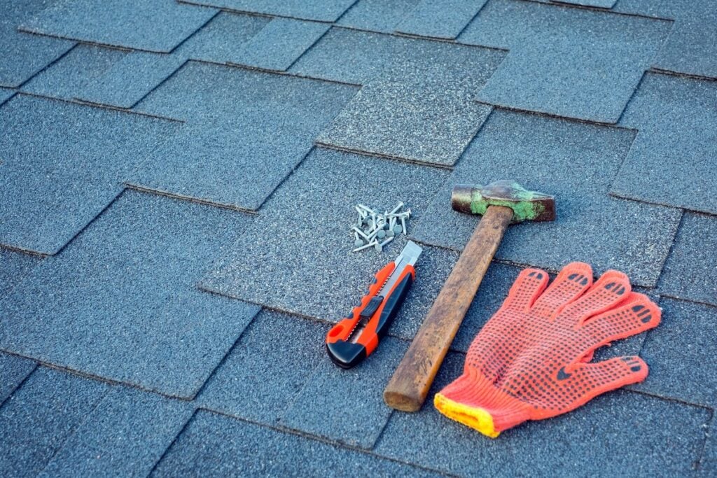 A utility knife, hammer, pile of nails, and an orange work glove—essential roofing tools—are laid out on gray asphalt roof shingles.