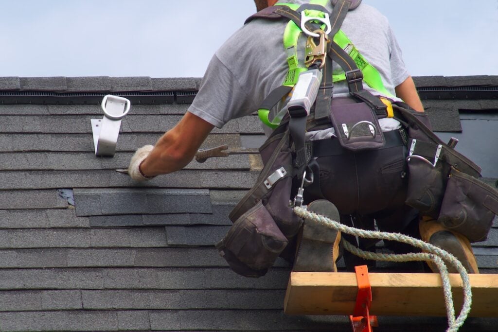 A worker wearing safety gear repairs asphalt roof shingles while standing on scaffolding. Using roofing tools, the worker secures materials around them, with a safety harness attached for fall protection. roofing tools