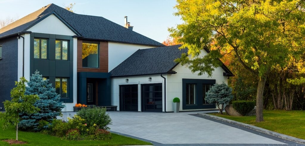 Modern two-story house with large windows, metal roofing, a double garage, and a clean driveway. The yard in Oakland, TN features manicured shrubs, trees, and green grass under sunlight filtering through the leaves.