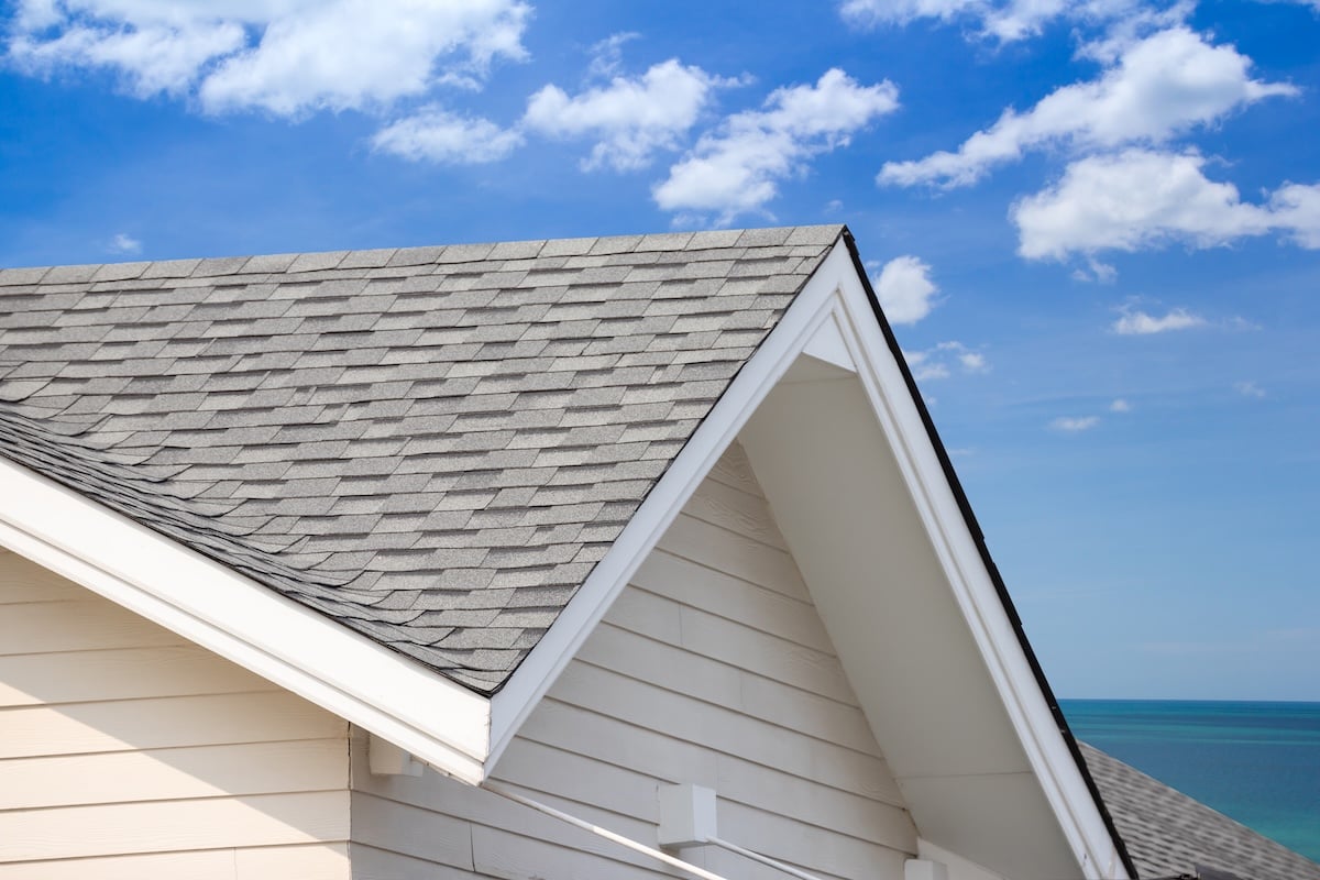 Close-up of a house roof with gray asphalt shingles and white siding, set against a blue sky with scattered clouds and a view of the ocean in the background, showcasing quality options like metal roofing in Oakland, TN.