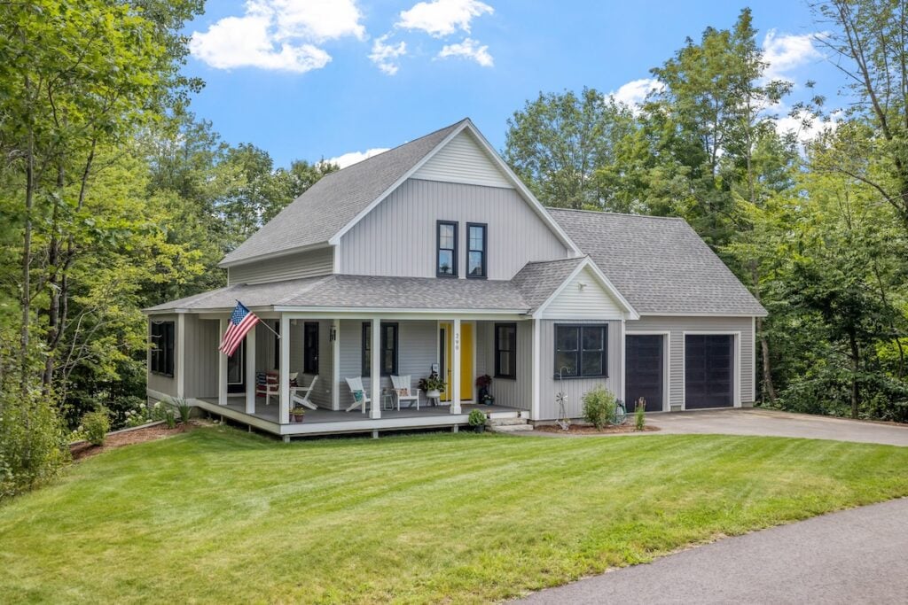 A modern two-story house with gray siding, a metal roofing accent, wraparound porch, black-framed windows, and a three-car garage sits amid lush green trees and a manicured lawn in Oakland TN. An American flag hangs by the porch.