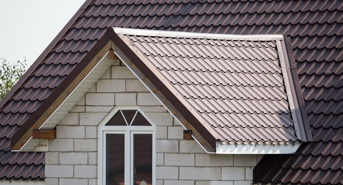 Close-up of a house roof with brown metal shingles, showcasing quality metal roofing Dickson TN, and a gable above a white-framed window set in a light-colored brick wall.