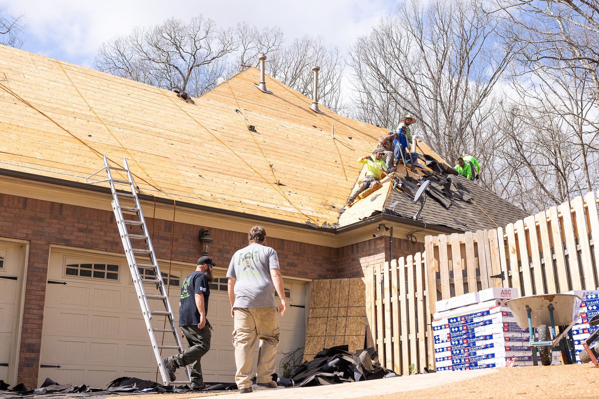 Several workers are installing new roofing on a house, with some on the roof and others on the ground. A tall ladder leans against the structure, and construction materials are stacked nearby. Leafless trees are visible in the background.