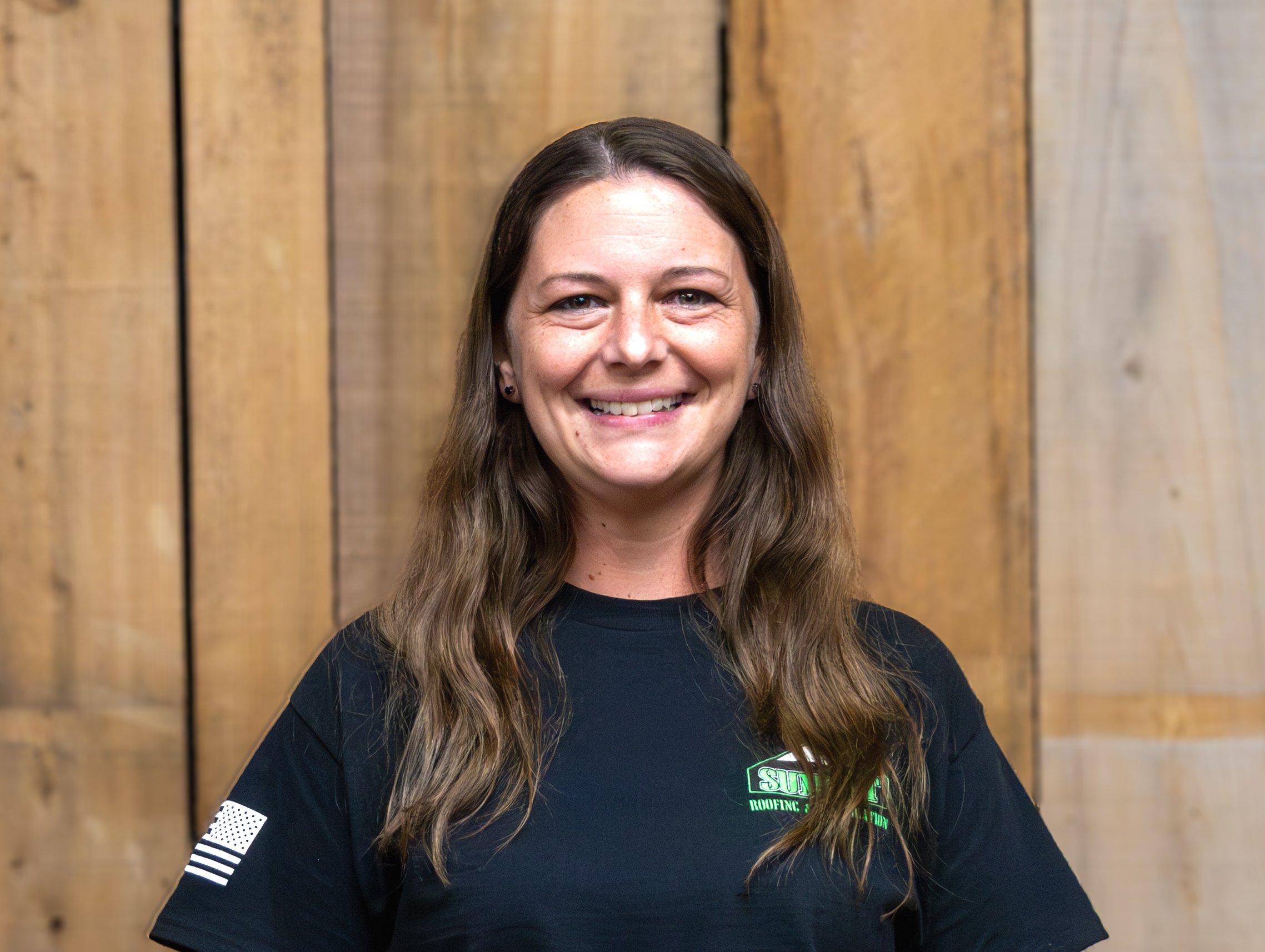 A woman with long brown hair, wearing a black T-shirt with a small green logo and an American flag on the sleeve, smiles while standing in front of a wooden wall.