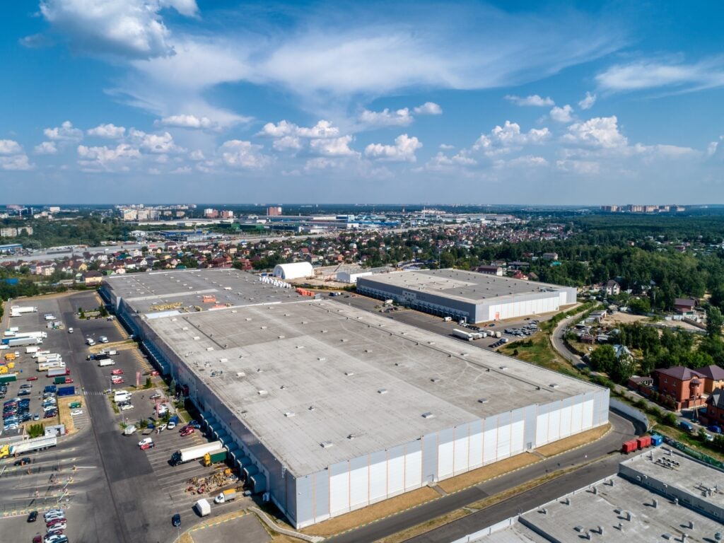 Aerial view of a large industrial warehouse complex featuring commercial flat roofing on multiple buildings, parking lots, and trucks, surrounded by greenery and a distant residential area under a blue sky with clouds.