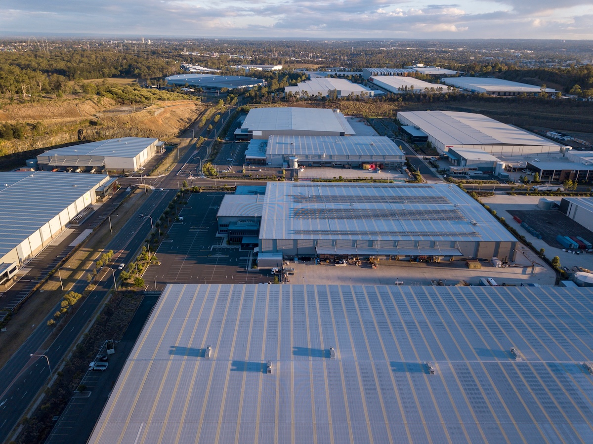 Aerial view of a large industrial area with wide warehouses featuring commercial roofing Pocahontas AR, solar panels on rooftops, parking lots, and roads, surrounded by greenery and distant cityscape under a partly cloudy sky.