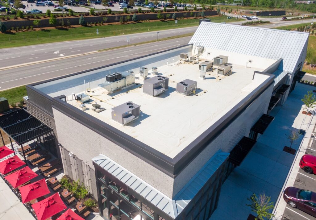 Aerial view of a commercial building with a flat white roof, multiple HVAC units, and a nearby road, showcasing expert commercial roofing Pocahontas AR. Red patio umbrellas and parked cars are visible at the lower left and right sides of the building.