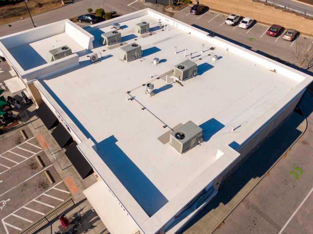 Aerial view of a commercial flat roofing structure with several HVAC units on its white roof, surrounded by a mostly empty parking lot with a few cars and clearly marked parking spaces. commercial flat roofing