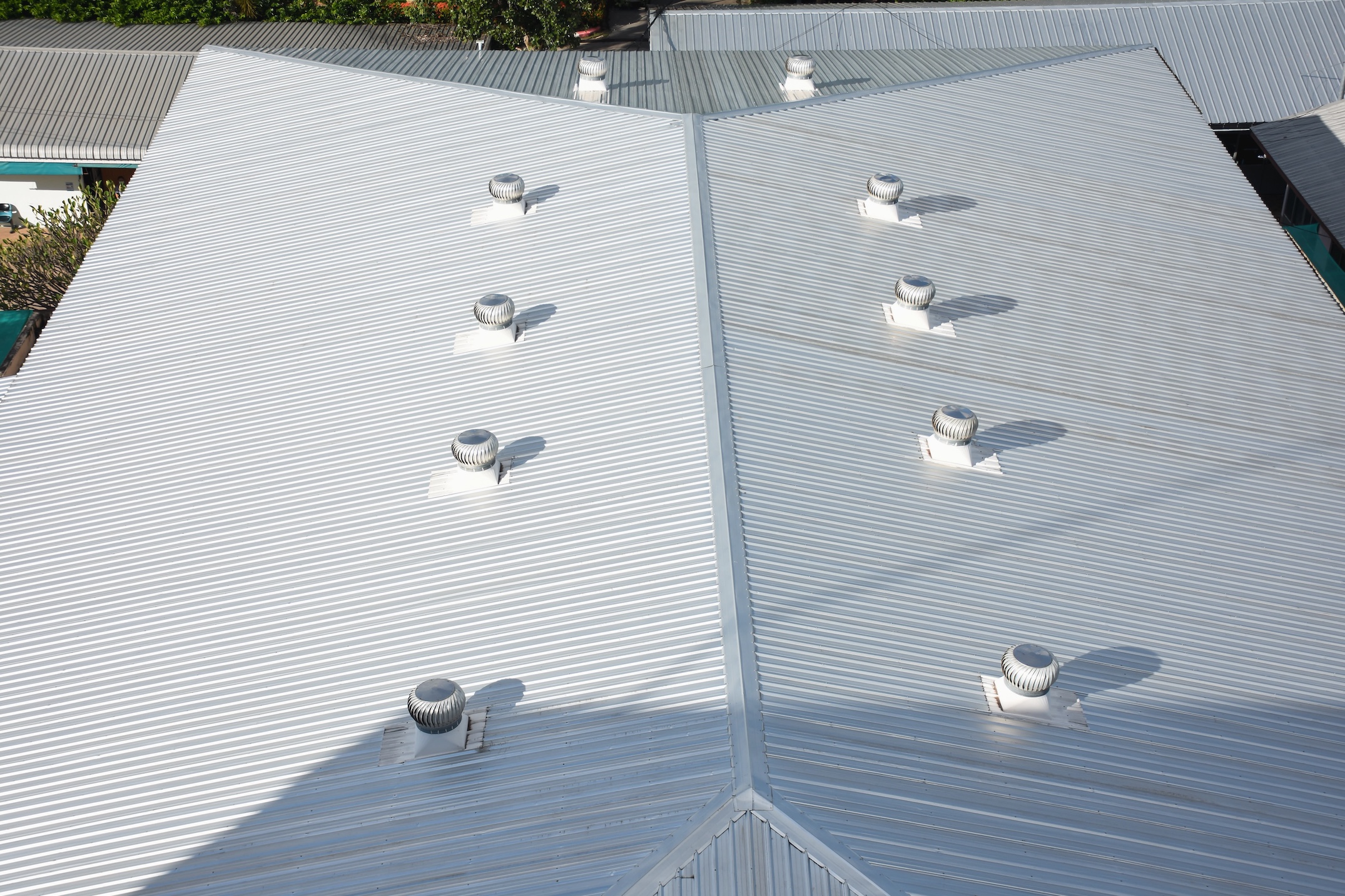 A top-down view of a large corrugated metal roof highlights its silver finish and several turbine vents, showcasing expert commercial roofing Germantown TN, all surrounded by greenery and neighboring buildings.
