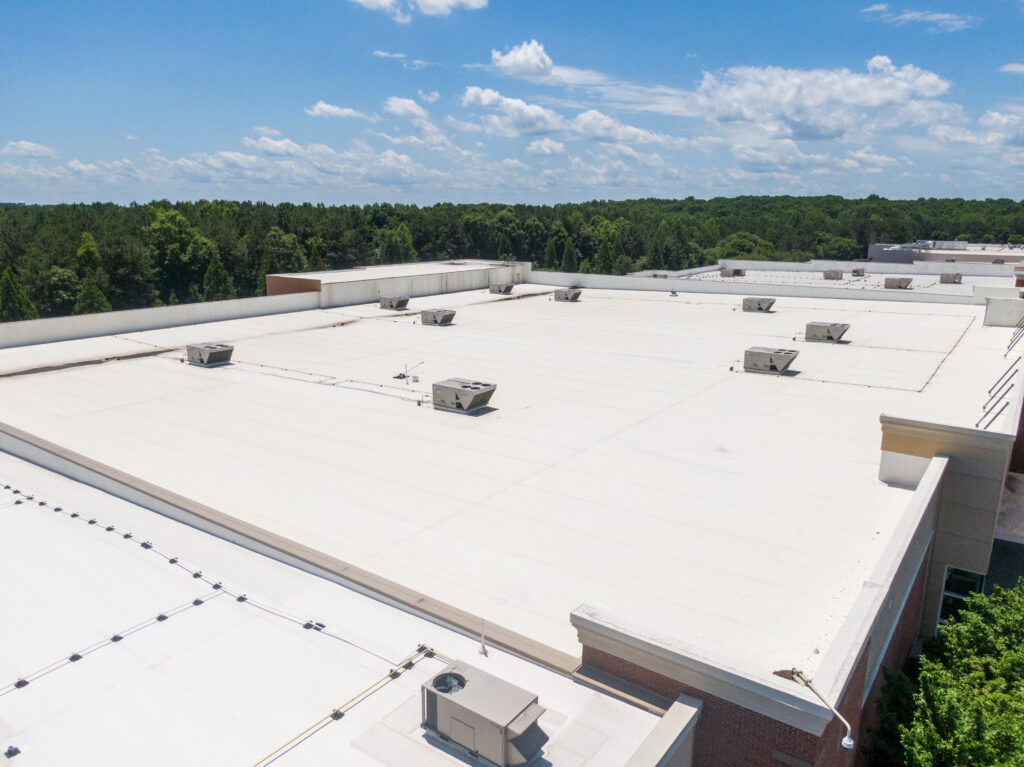 Aerial view of a large, flat white commercial roofing Germantown TN rooftop with multiple HVAC units, surrounded by trees under a blue sky with scattered clouds.