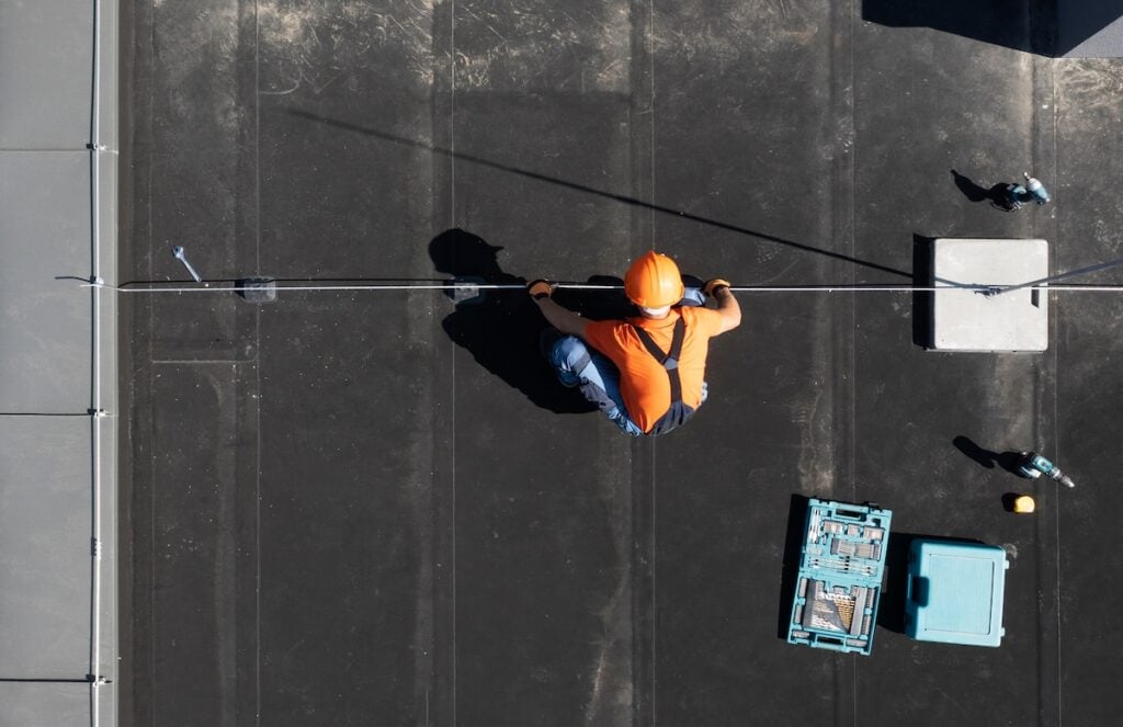 A worker wearing an orange helmet and vest kneels on a commercial flat roofing surface, handling a cable. A blue tool box and various tools are nearby. The scene is viewed from directly above.