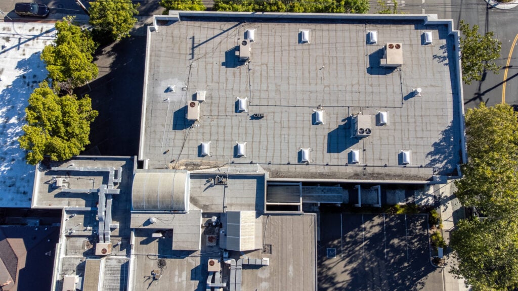 Aerial view of a commercial flat roofing building surrounded by trees and roads, with several vents, air conditioning units, and utility structures visible on the rooftop.