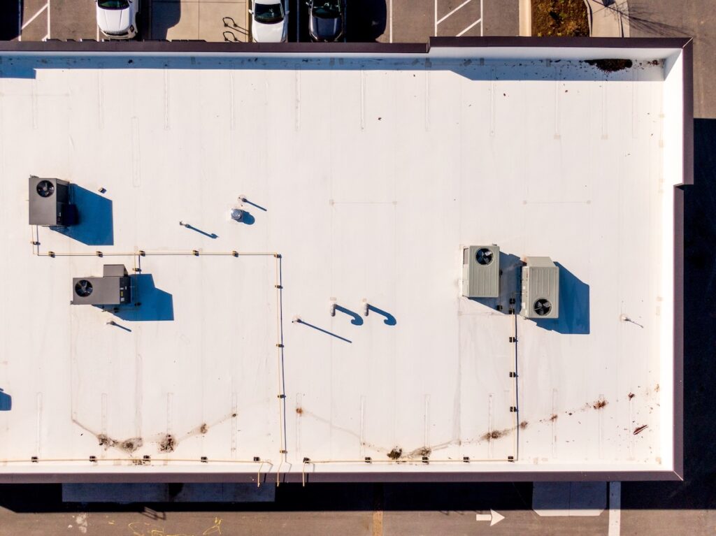 Aerial view of a flat, white commercial roofing Paragould AR building with HVAC units, pipes, and vents. Some dirt and debris are scattered near the roof edges. Cars are parked along the top side of the image.