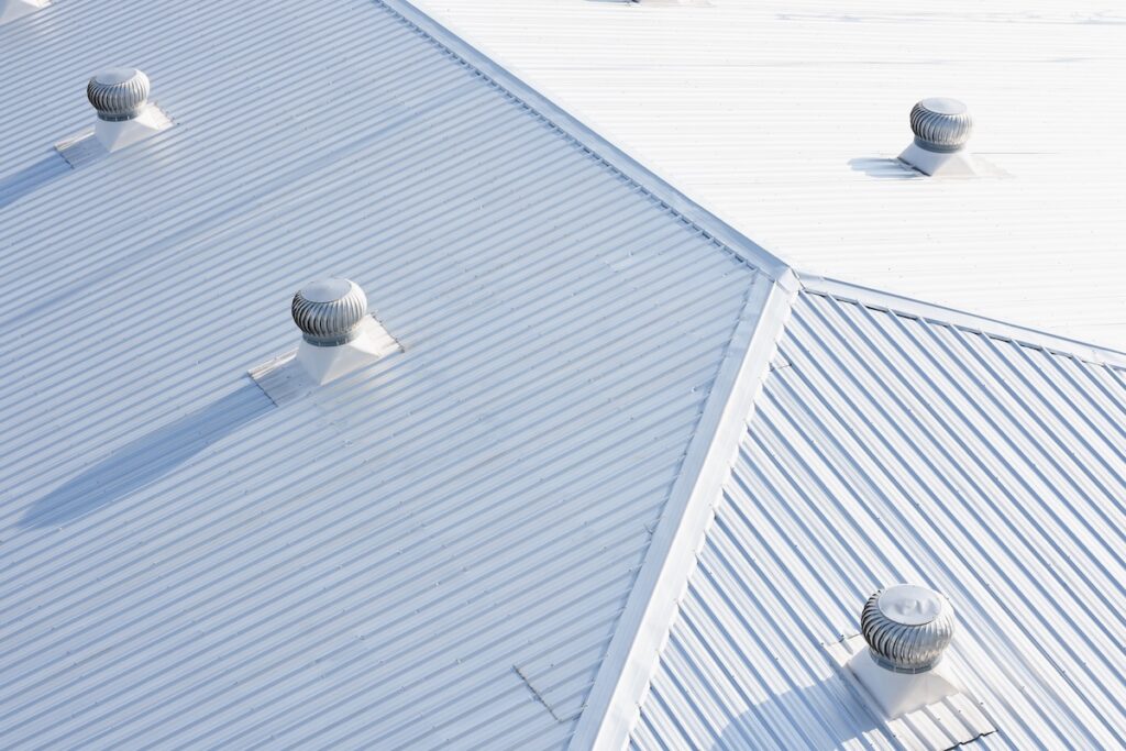 Overlapping white metal roofs with ridged patterns and several silver turbine roof vents casting long shadows in bright sunlight, showcasing expert commercial roofing Paragould AR.