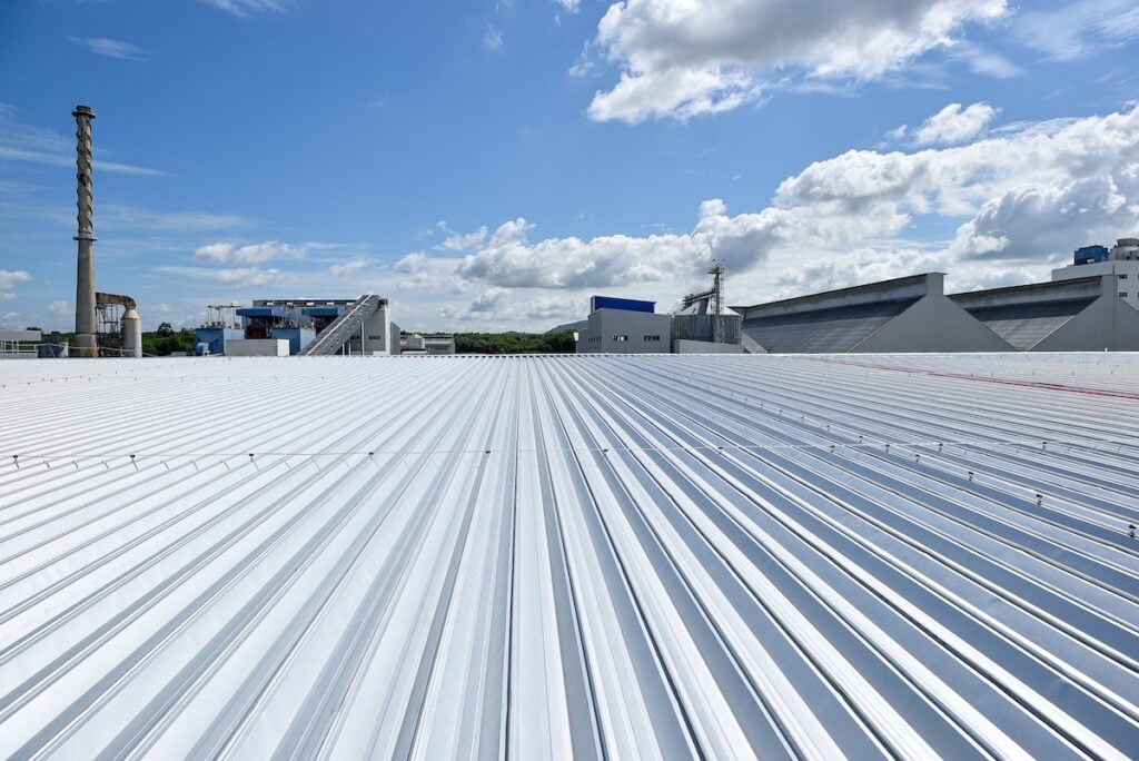 A wide, bright metal roof extends into the distance under a blue sky with scattered clouds; commercial roofing in Waverly, TN stands out, with industrial buildings and chimneys visible on the horizon.