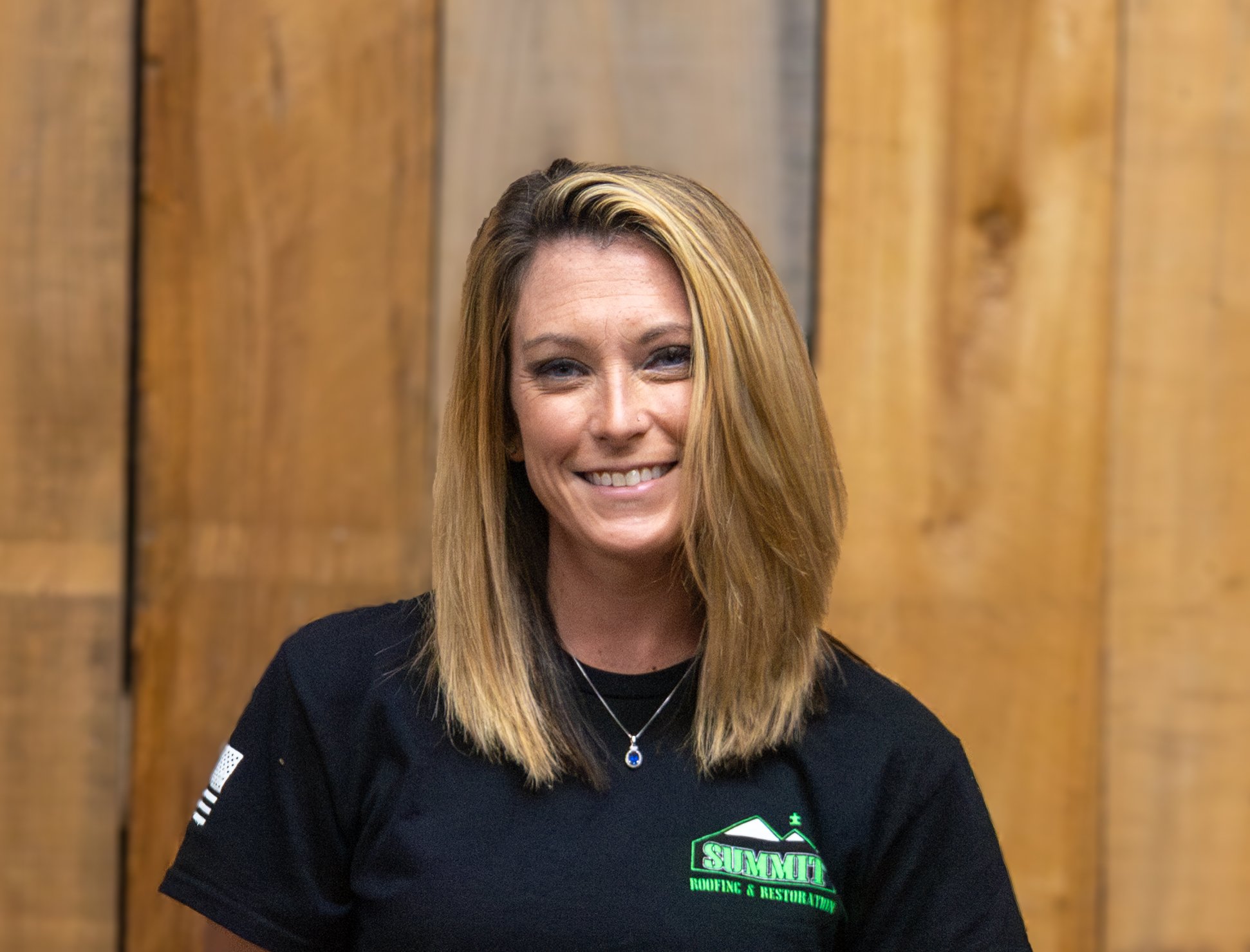A woman with straight, shoulder-length blonde hair smiles at the camera. She is wearing a black Summit Roofing & Restoration t-shirt and a necklace, standing in front of a wooden plank background.