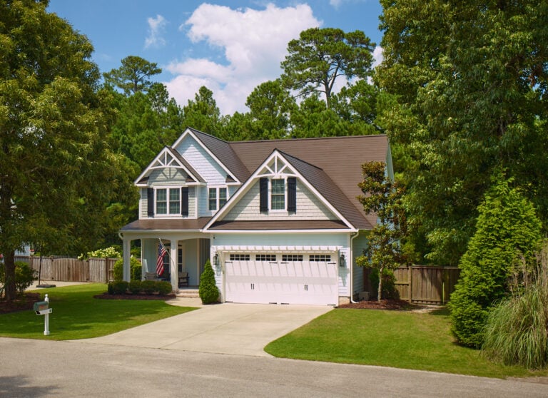 A two-story suburban house with a gabled roof, light gray siding, and white trim. The home has a two-car garage, a front porch with an American flag, and is surrounded by trees and a neatly kept lawn.