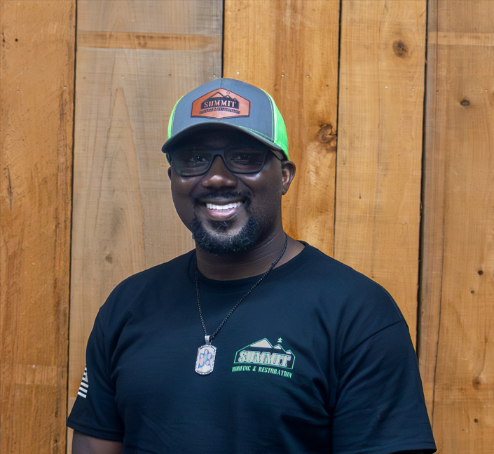 A man wearing glasses, a gray and green Summit cap, and a black Summit Home & Restoration shirt stands smiling in front of a wooden wall.