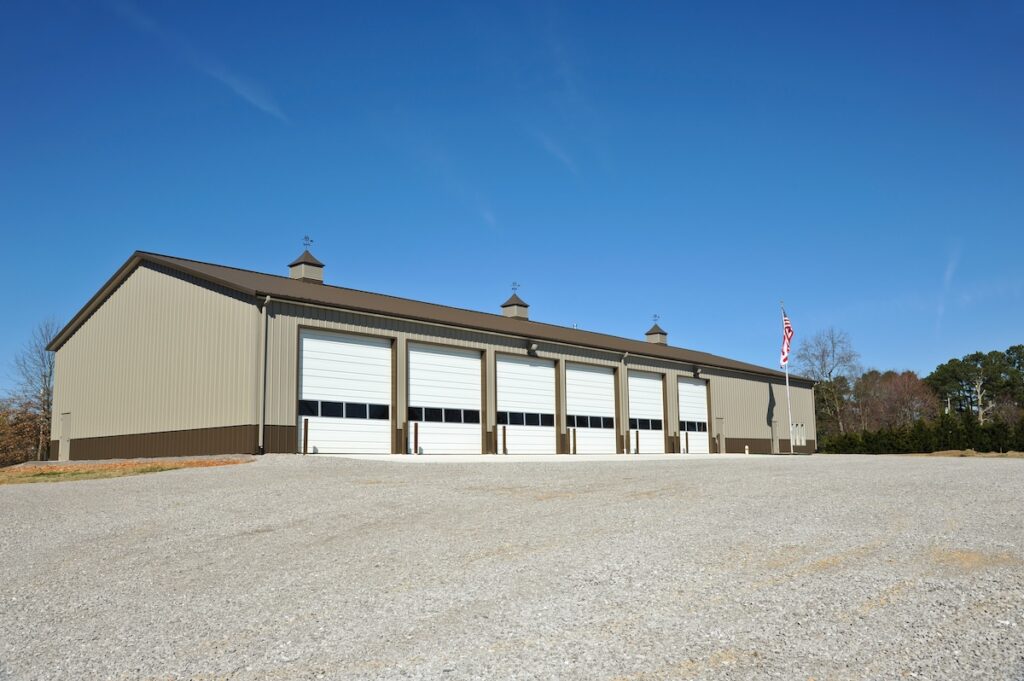 A large, tan metal building with five white garage doors stands on a gravel lot under a clear blue sky. An American flag waves on a pole to the right, with trees behind—ideal for commercial roofing in Caruthersville MO.