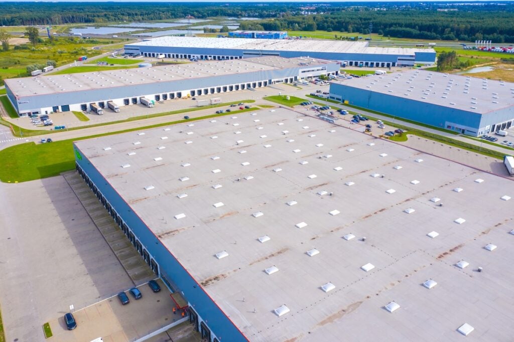 Aerial view of a large industrial complex with multiple expansive warehouse buildings, surrounding parking lots with cars, and green fields in the background under a partly cloudy sky. flat roof maintenance