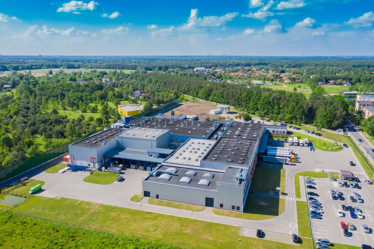 Aerial view of a large industrial building or factory complex with commercial roofing in Clarksville, TN, surrounded by greenery, trees, and open fields, with a parking lot and vehicles near the entrance under a blue sky with scattered clouds.