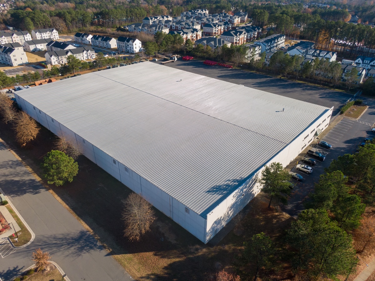 A large white warehouse with a metal roof, showcasing expert commercial roofing in Caruthersville, MO, is surrounded by a parking lot with several cars. Apartment buildings and trees are visible in the background on a clear day.