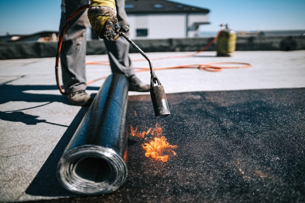 A worker uses a blowtorch to heat and install roofing material on a flat rooftop. A roll of roofing membrane is partially unrolled, and protective gloves cover the worker's hands.