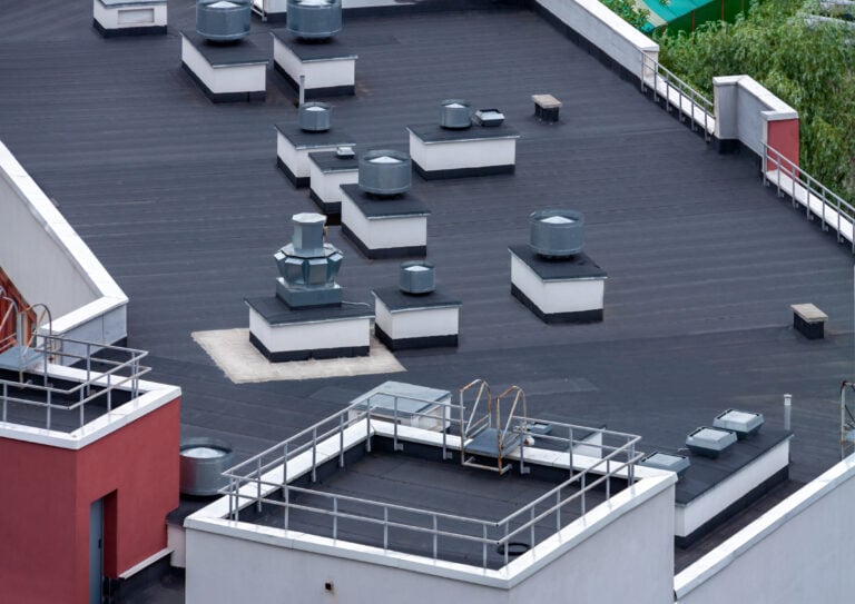 A flat rooftop with multiple ventilation units, ducts, and skylights highlights the importance of flat roof maintenance. The black surface is bordered by white and red walls, railings, and a ladder for access. Green trees appear in the background.