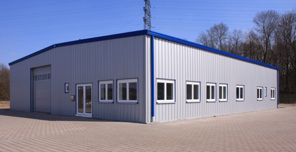A large, gray industrial warehouse with multiple windows and a blue roof trim showcases quality commercial roofing Clarksville TN; it's set on a paved lot with trees and power lines in the background under a clear sky.
