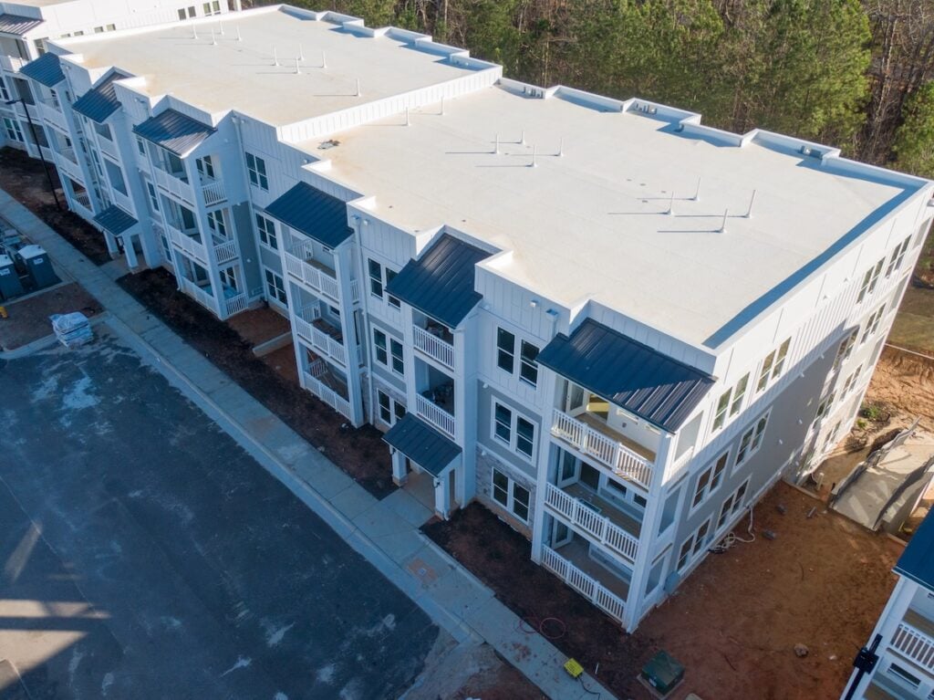 Aerial view of a newly constructed three-story apartment building with white exterior, commercial roofing Waverly TN, balconies, and an adjacent paved parking area, surrounded by trees and exposed soil.
