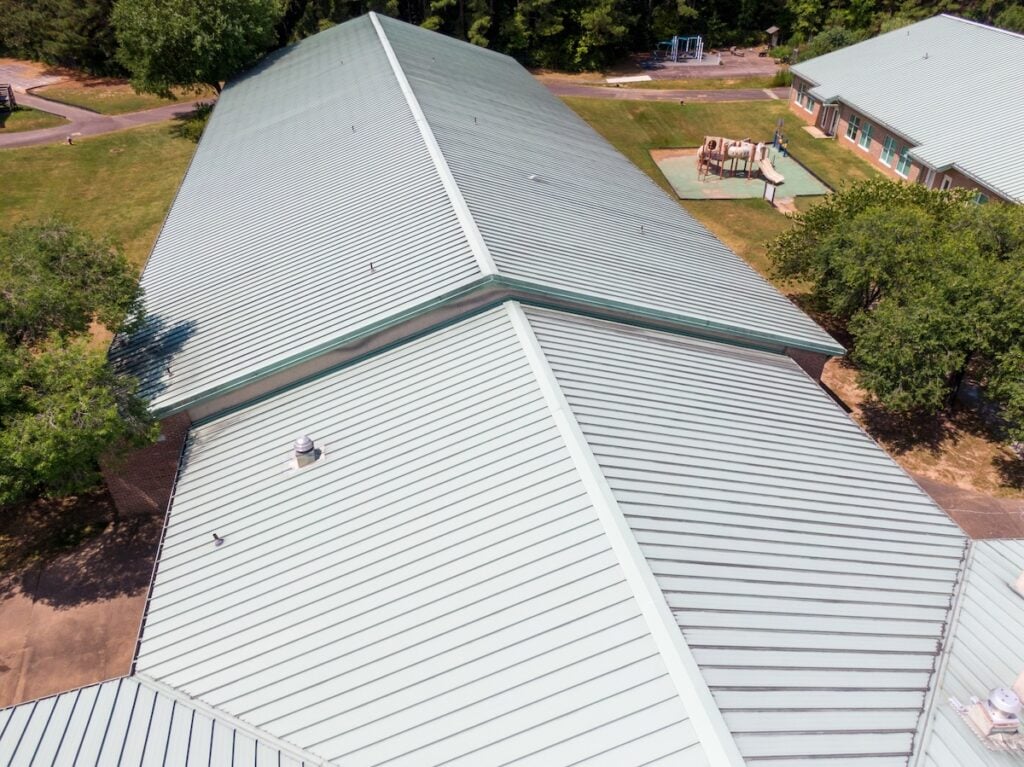 Aerial view of a large, light green metal roof on a building surrounded by grass, trees, and paved walkways in Milan, TN—showcasing quality commercial roofing with a playground visible in the background.
