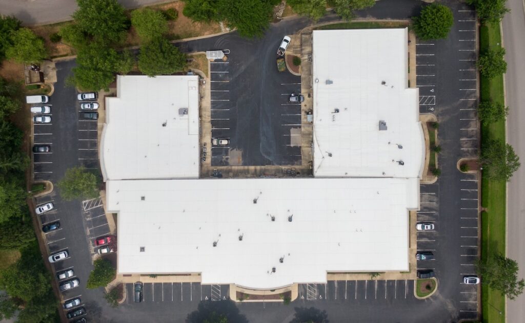 Aerial view of a large, white commercial roofing Caruthersville MO building with a central parking lot surrounded by trees and parked cars. The C-shaped structure is bordered by roads on three sides.