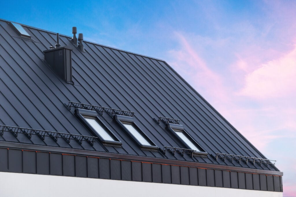 A modern, dark metal mansard roof with skylights and safety railings, set against a blue sky with light pink clouds.