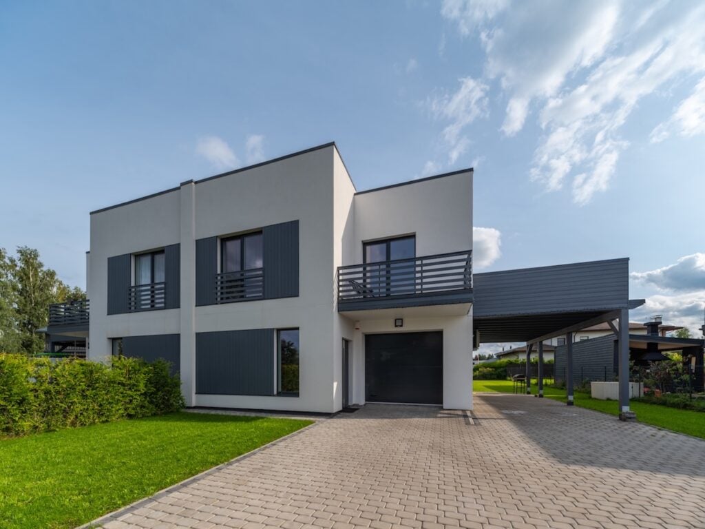 Modern two-story house with a white facade, large windows, gray shutters, and a stylish flat roof. Features include a driveway, carport, green lawn, hedge, and a blue sky with scattered clouds.
