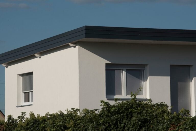 A modern white house with a flat roof features two windows with closed blinds and green shrubbery in front, all set under a clear blue sky.