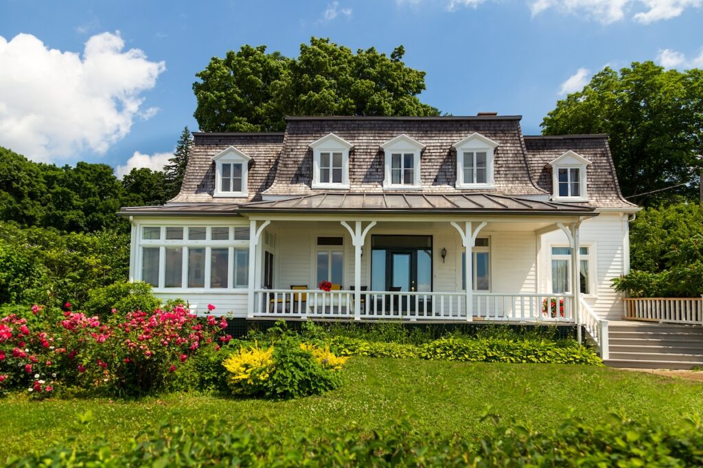 Charming white house with a mansard roof, wide front porch, and dormer windows. Bright flowers and shrubs border the house, with large trees and a blue sky in the background.
