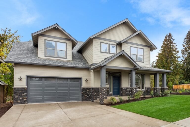 Modern two-story house with beige siding, gable roof, and stone accents. Features a double garage, large windows, a covered front porch, and a well-maintained lawn with trees in the background under a bright blue sky.