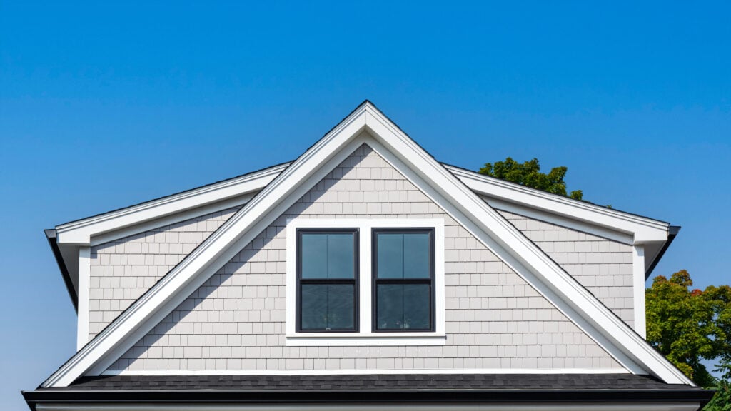A close-up view of a gable roof with gray shingle siding, two black-framed windows, and a clear blue sky in the background. Green tree branches are visible on the right side. 