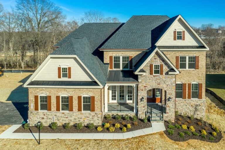 A two-story suburban house with a stone and brick exterior, multiple gables including a charming gambrel roof, shuttered windows, and a landscaped front yard with bushes and a curved walkway under a clear blue sky.