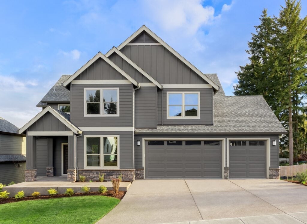 Modern two-story house with a gable roof, gray siding, large windows, and a three-car garage. The front yard has a manicured lawn, small shrubs, and a wide driveway. Tall trees are visible in the background under a blue sky. gable roof