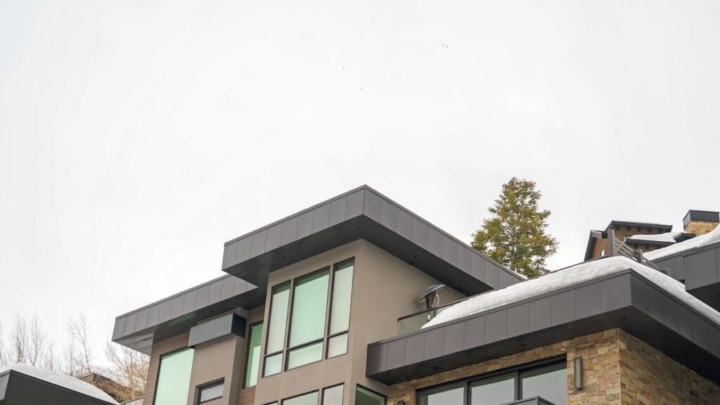Modern house with large windows and a flat roof, partially covered with snow. The building showcases stone accents and dark trim, while trees rise against a cloudy sky in the background. flat roof