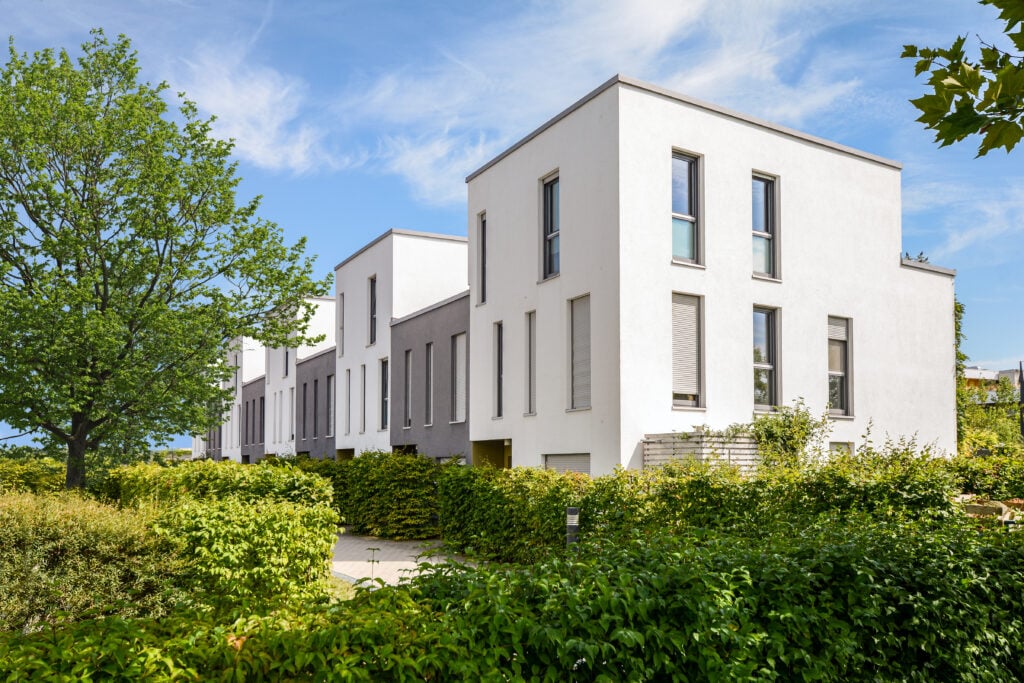 Row of modern white townhouses with large windows and stylish flat roofs, bordered by neatly trimmed green hedges, all set under a blue sky with scattered clouds. flat roof