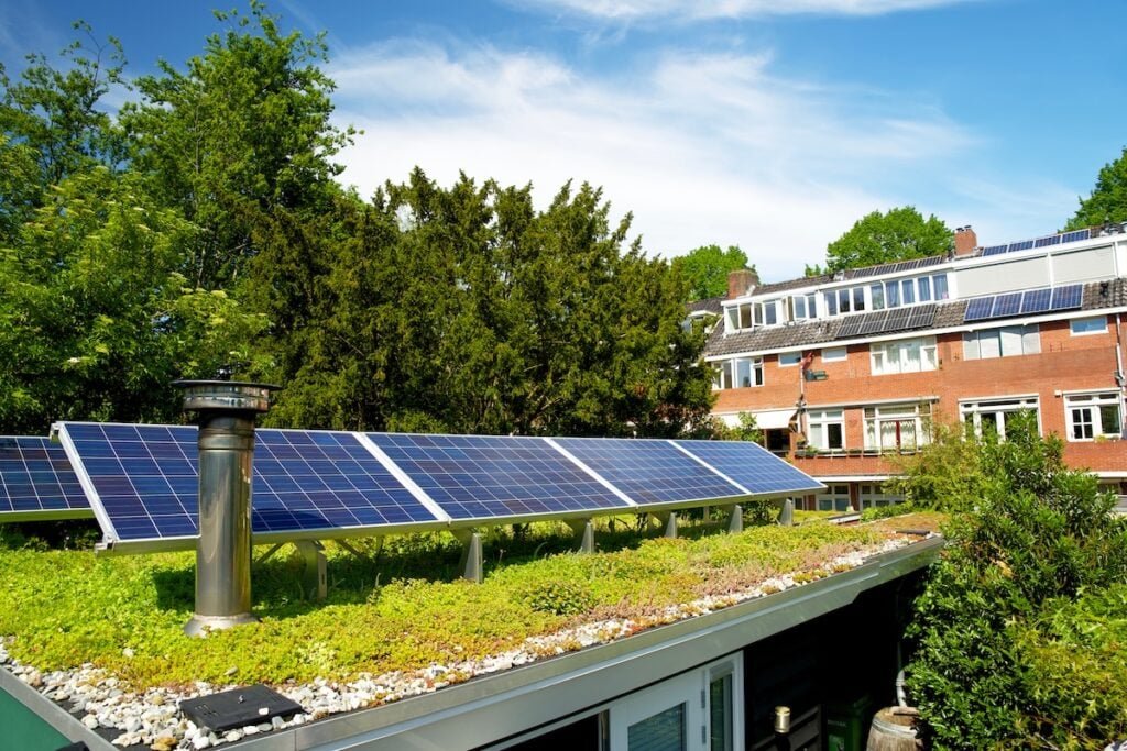 Solar panels are installed on the building’s green roof, surrounded by lush plants and trees. More solar panels can be seen atop neighboring residential buildings, all set beneath a clear blue sky.