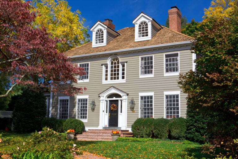 A classic two-story colonial-style house with gray siding, white trim, and dormer windows, surrounded by autumn trees and neatly trimmed bushes. The front door is decorated with a fall wreath.
