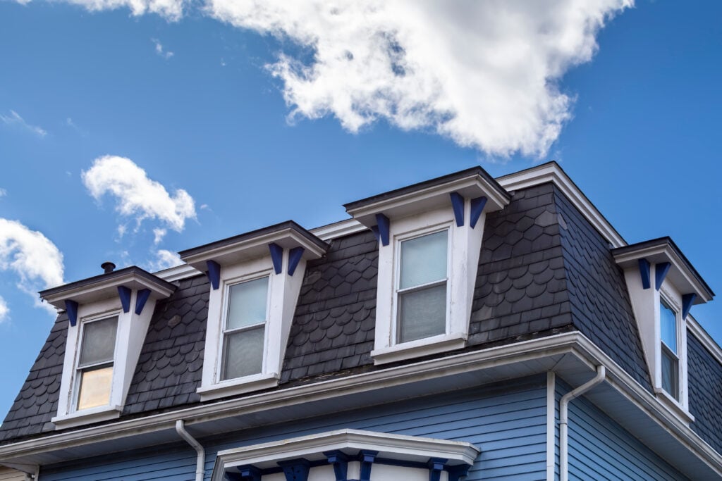 The image shows the top part of a blue house with three dormer windows, dark gray fish-scale shingles, and white trim, set against a bright blue sky with scattered clouds.