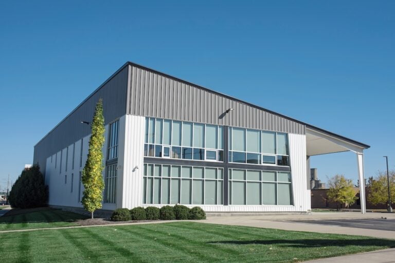 A modern industrial or commercial building with a sleek shed roof, large glass windows, and metal siding, surrounded by neatly trimmed grass, bushes, and a few trees under a clear blue sky.