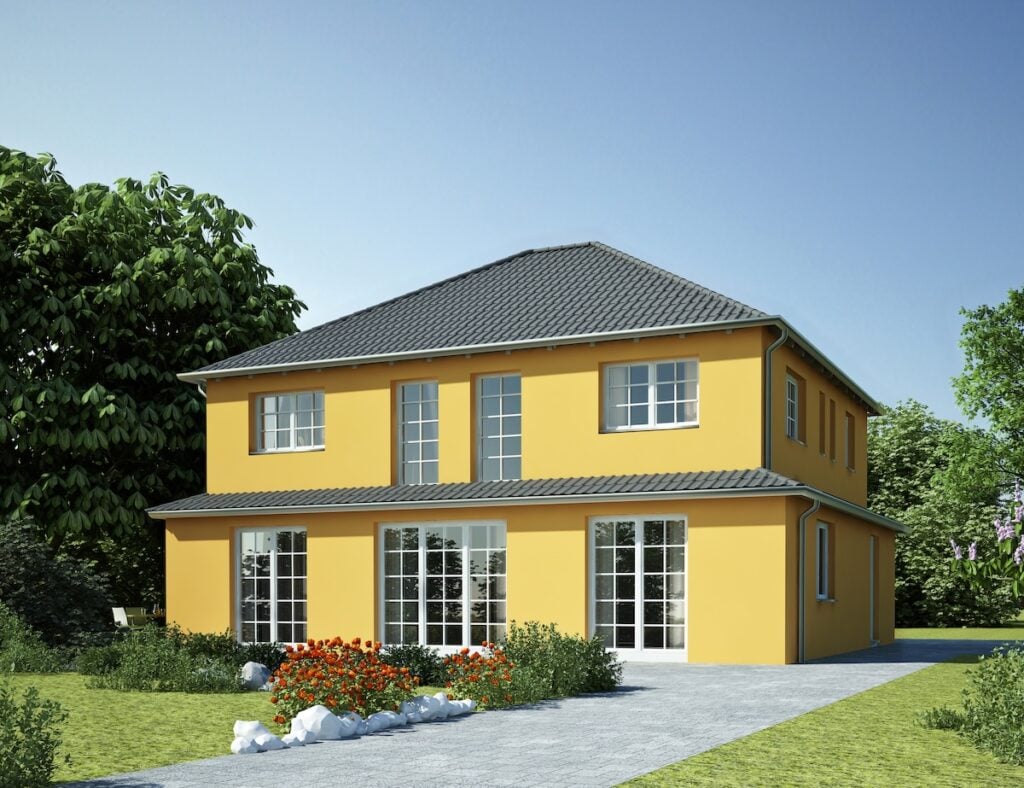 A modern two-story yellow house with large windows, a gray tiled roof, and a garden featuring flowers and greenery along a stone walkway, with trees in the background under a clear blue sky.