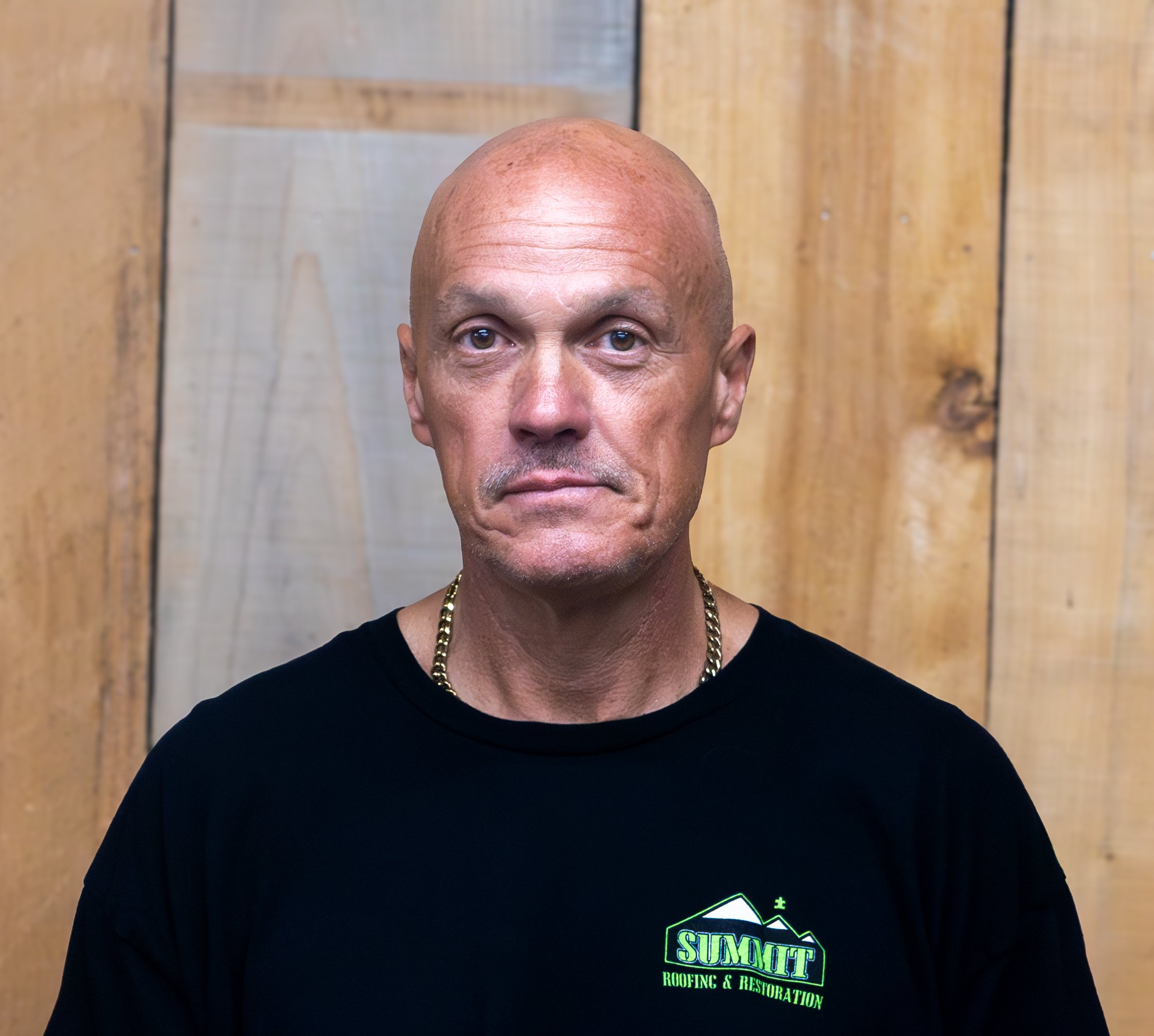 A middle-aged man with a shaved head and gold chain wears a black